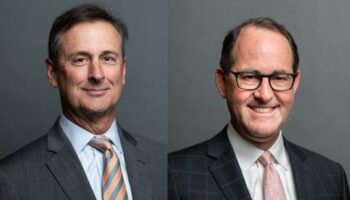 Two Men In Business Suits Pose Against A Plain Gray Background. The Man On The Left Wears A Gray Suit And Striped Tie; The Man On The Right Wears Glasses, A Checkered Suit, And A Light Pink Tie. Both Are Smiling.