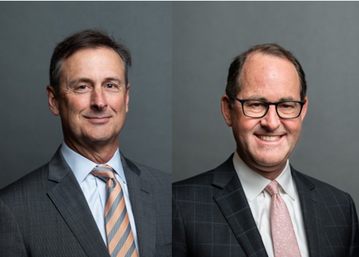 Two men in business suits pose against a plain gray background. The man on the left wears a gray suit and striped tie; the man on the right wears glasses, a checkered suit, and a light pink tie. Both are smiling.