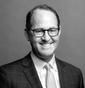 Black and white portrait of a man wearing glasses, a suit, and a tie. He is smiling and facing the camera, with a plain background behind him.