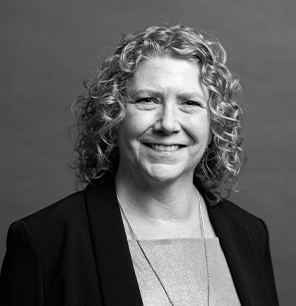 Black and white portrait of a smiling woman with curly hair, wearing a blazer over a light-colored top and a necklace, posed in front of a plain background.