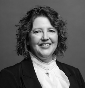 Black and white portrait of a woman with wavy hair, wearing a dark blazer over a ruffled white blouse, smiling at the camera against a plain background.