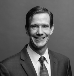 A man in a suit and tie smiles at the camera against a plain background in a black-and-white portrait.