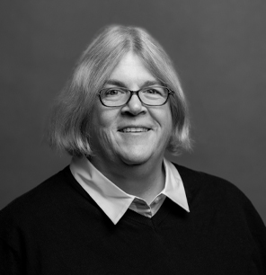 A person with shoulder-length light hair, glasses, and a friendly smile is wearing a collared shirt under a dark sweater, posing against a plain, neutral background.