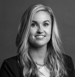 Black and white portrait of a woman with long, wavy blonde hair, wearing a dark blazer, light top, and a chain necklace, smiling at the camera against a plain background.