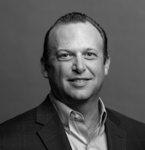 Black and white portrait of a man with short, slicked-back hair, wearing a suit jacket and collared shirt, smiling at the camera against a plain background.