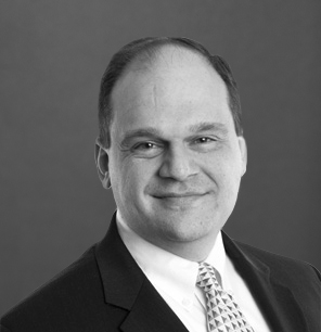 A man with short hair wearing a suit, white shirt, and patterned tie is smiling slightly. The photo is in black and white with a plain background.