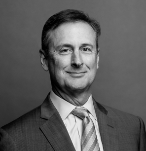 A middle-aged man in a suit and striped tie poses against a plain background, looking at the camera with a slight smile. The image is in black and white.