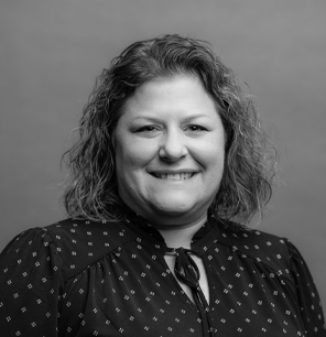 Black and white portrait of a woman with curly shoulder-length hair, wearing a patterned blouse with a tie at the collar, smiling in front of a plain background.