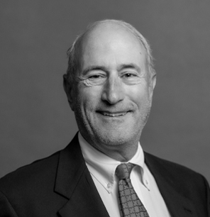 A smiling older man with short, thinning hair wears a suit jacket, white dress shirt, and patterned tie, posing in front of a plain, neutral background. The image is in black and white.