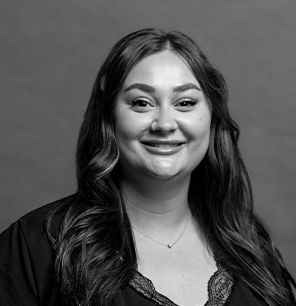 Black and white portrait of a woman with long, wavy hair. She is smiling, wearing a necklace and a dark top, and the background is plain and neutral.