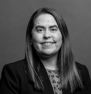 Black and white portrait of a woman with long straight hair, wearing a blazer over a patterned top, smiling at the camera against a plain background.