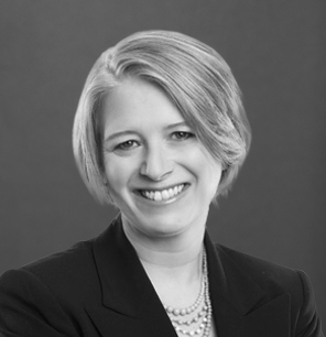 Black and white portrait of a woman with short, light-colored hair, wearing a dark blazer and a pearl necklace, smiling at the camera against a plain background.