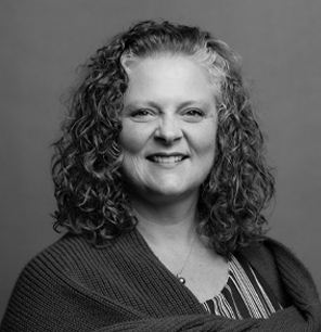 Black and white portrait of a woman with curly hair, wearing a striped top and a textured shawl, smiling softly at the camera against a plain background.