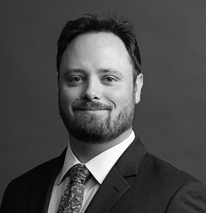 A man with short dark hair and a beard, wearing a suit, patterned tie, and white shirt, smiles slightly in a professional black-and-white portrait against a plain background.