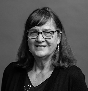 A woman with shoulder-length hair and glasses smiles at the camera. She is wearing earrings, a dark top, and a cardigan. The background is plain and out of focus. The photo is in black and white.