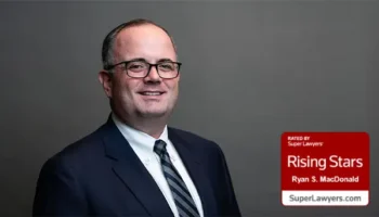 A Man In A Dark Suit, White Shirt, And Striped Tie Smiles At The Camera Against A Gray Background. A Red Badge In The Corner Reads “Rising Stars – Ryan S. MacDonald – SuperLawyers.com.”.