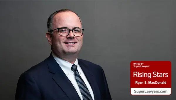 A man in a dark suit, white shirt, and striped tie smiles at the camera against a gray background. A red badge in the corner reads “Rising Stars – Ryan S. MacDonald – SuperLawyers.com.”.