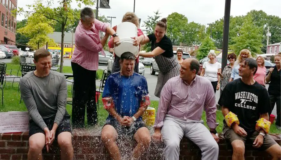 A group of people outside watch as a man seated on a low brick wall gets a bucket of ice water dumped on him, splashing water everywhere; three other men sit beside him, some wearing water wings.