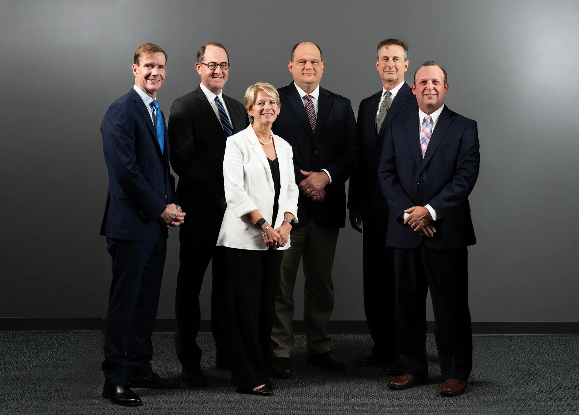 Six professionally dressed adults, five men in dark suits and one woman in a white blazer, stand posing and smiling in front of a plain gray background.