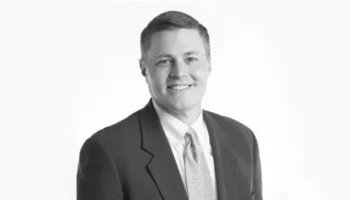 A Man In A Suit And Tie Smiles At The Camera Against A Plain, Light Background. The Image Is In Black And White.