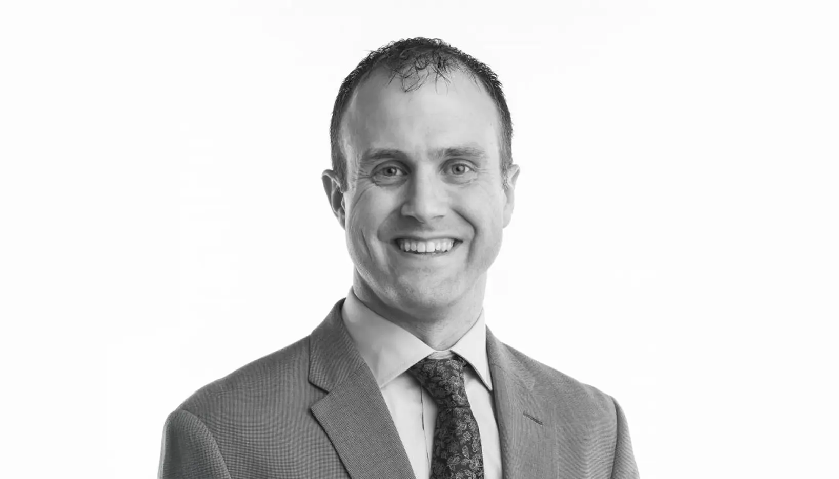 A man in a suit and tie smiles at the camera against a plain, light background. The image is in black and white.