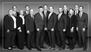 A Group Of Thirteen Professionally Dressed Adults, Including Men In Suits And Women In Business Attire, Standing Together And Smiling For A Formal Group Photo Against A Plain Background.