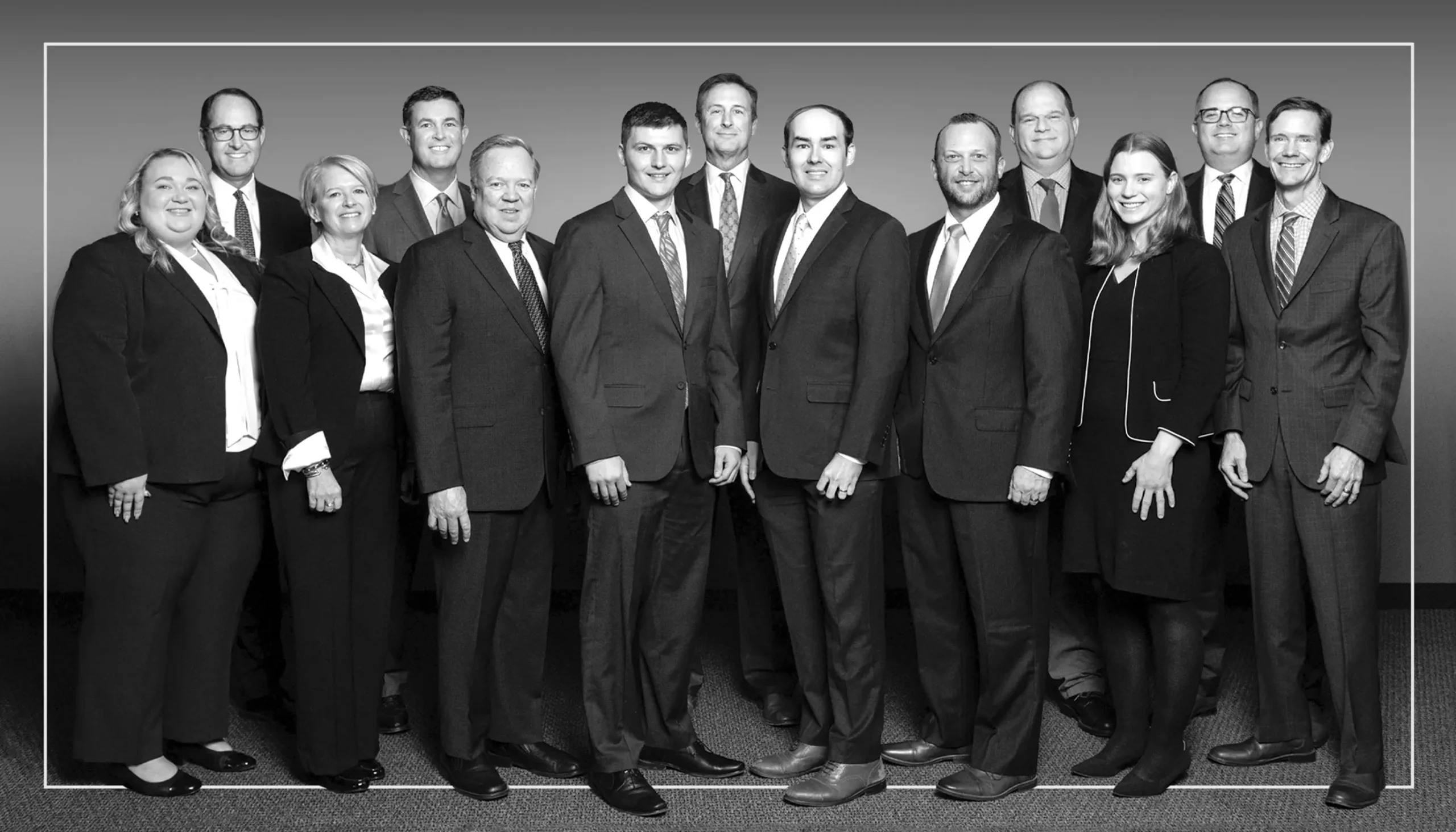 A group of thirteen professionally dressed adults, including men in suits and women in business attire, standing together and smiling for a formal group photo against a plain background.
