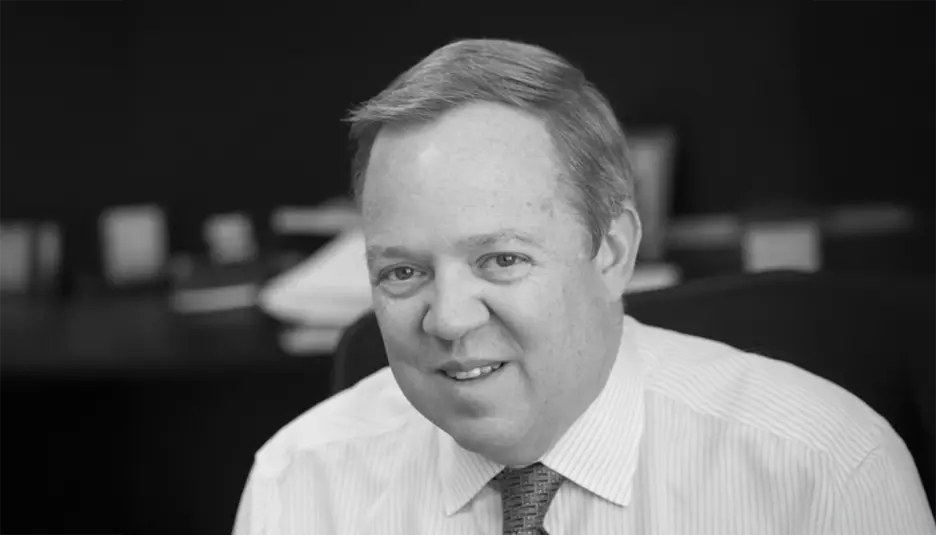 A man in a dress shirt and tie sits in an office, smiling at the camera. The background is blurred, and the image is in black and white.