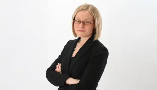 A woman with straight blonde hair and glasses stands with arms crossed, wearing a black suit jacket against a plain white background.
