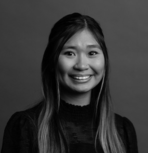 Black and white portrait of a young woman with long, straight hair, wearing a textured dark top, smiling and looking at the camera against a plain background.