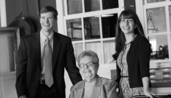 Three People Pose Together Indoors: An Older Woman Sitting And Smiling, With A Man In A Suit And A Woman In A Dress Standing Beside Her. All Three Are Looking Toward The Camera And Smiling. The Image Is In Black And White.