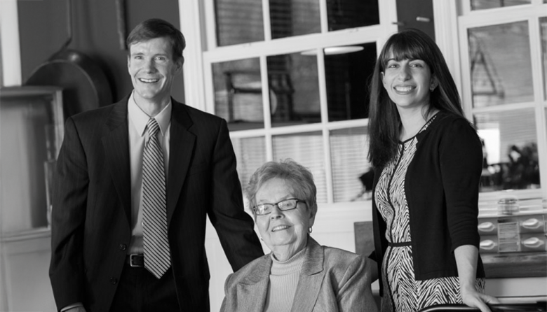 Three people pose together indoors: an older woman sitting and smiling, with a man in a suit and a woman in a dress standing beside her. All three are looking toward the camera and smiling. The image is in black and white.