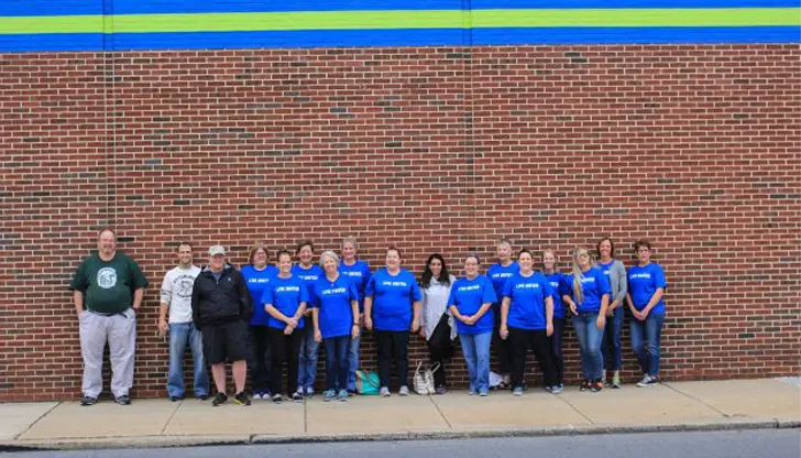 A group of people stand in a line against a brick wall. Most are wearing matching blue shirts, while a few wear different colors. The wall has a blue and green stripe near the top. The group stands on a sidewalk.