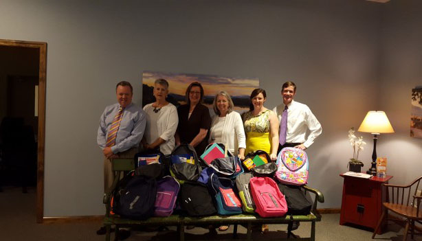 Six adults stand behind a bench filled with colorful backpacks and school supplies in a warmly lit room with blue walls, a lamp, and a table in the background.