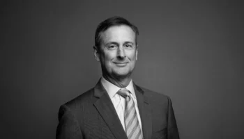 A Middle-aged Man In A Suit And Tie Smiles Slightly While Facing The Camera, Set Against A Plain, Dark Background. The Photo Is In Black And White.