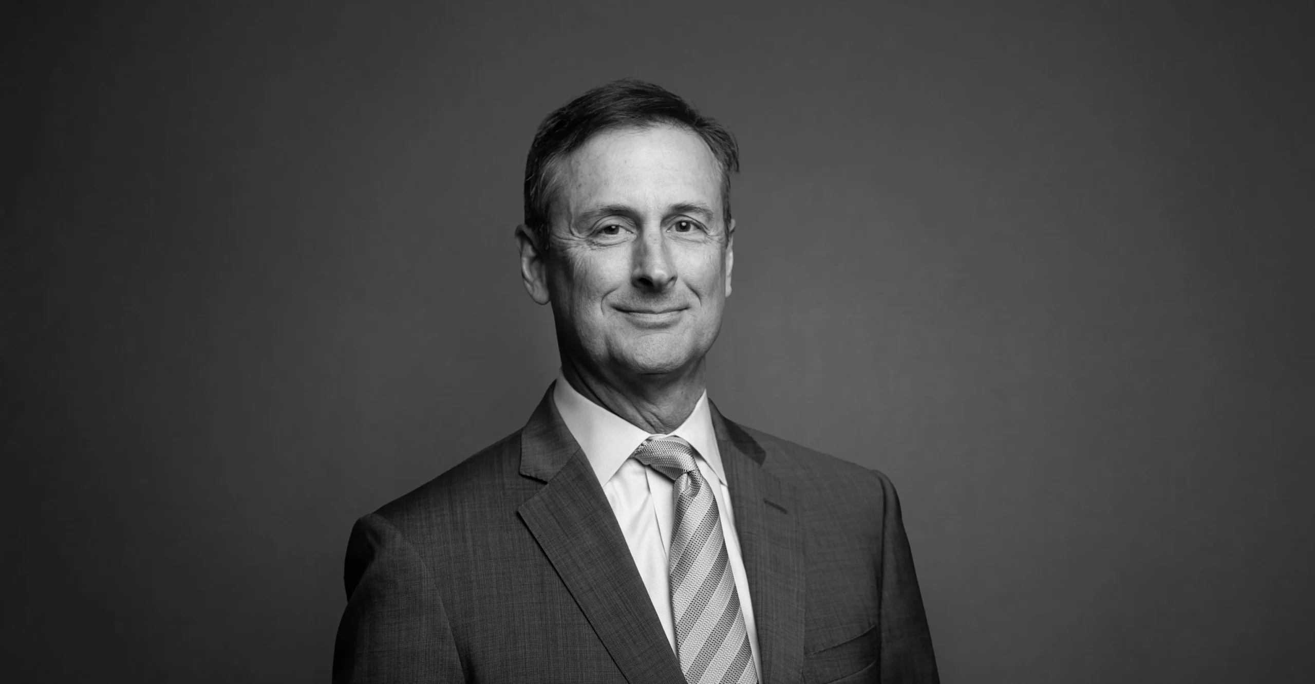 A middle-aged man in a suit and tie smiles slightly while facing the camera, set against a plain, dark background. The photo is in black and white.