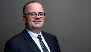 A Man Wearing Glasses, A Dark Suit, A White Dress Shirt, And A Striped Tie Stands Smiling Against A Plain Gray Background.