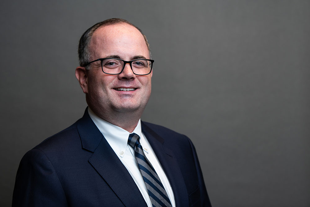 A man wearing glasses, a dark suit, a white dress shirt, and a striped tie stands smiling against a plain gray background.