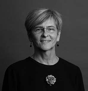 Black and white portrait of an older woman with short, light hair, wearing glasses, earrings, and a dark top with a decorative flower pin. She is smiling softly and facing the camera against a plain background.