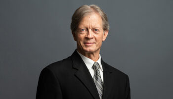 A Middle-aged Man With Light Skin And Gray Hair, Wearing A Black Suit, White Shirt, And Striped Tie, Stands Against A Plain Gray Background, Looking Slightly To The Side.