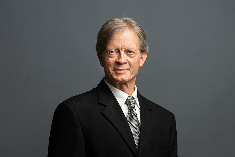 A middle-aged man with light skin and gray hair, wearing a black suit, white shirt, and striped tie, stands against a plain gray background, looking slightly to the side.