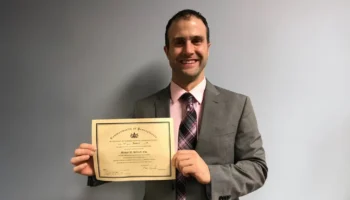 A Man In A Gray Suit And Pink Shirt Stands Against A Plain Wall, Smiling And Holding Up A Certificate With Both Hands.
