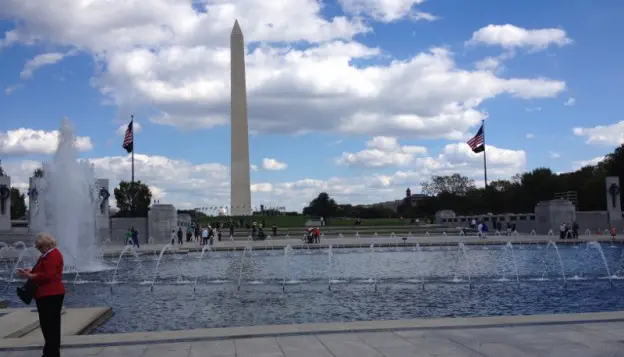 The Washington Monument stands tall under a partly cloudy sky, with American flags and fountains in the foreground and people walking around the National Mall in Washington, D.C.