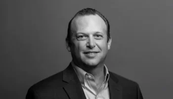 A Man In A Suit And Collared Shirt Smiles At The Camera In A Black And White Portrait With A Plain Background.