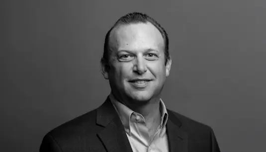 A man in a suit and collared shirt smiles at the camera in a black and white portrait with a plain background.