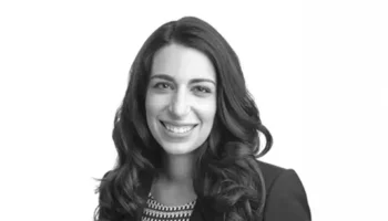 A Woman With Long, Wavy Dark Hair Is Smiling At The Camera. She Is Wearing A Patterned Top And A Dark Blazer. The Background Is Plain White.