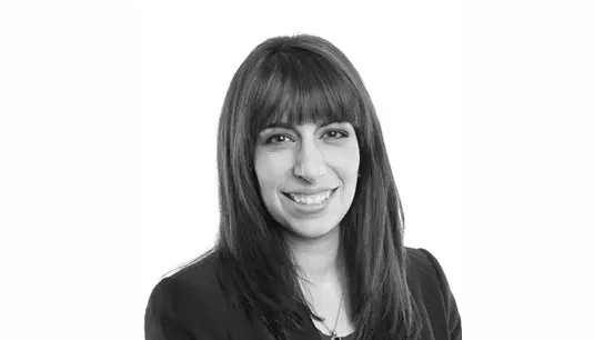 A woman with straight dark hair and bangs, wearing a dark blazer, smiles at the camera against a plain white background.
