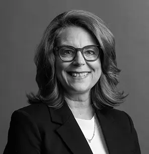 A woman with shoulder-length wavy hair and glasses smiles at the camera. She is wearing a dark blazer over a light top and a necklace. The background is plain and gray. The image is in black and white.