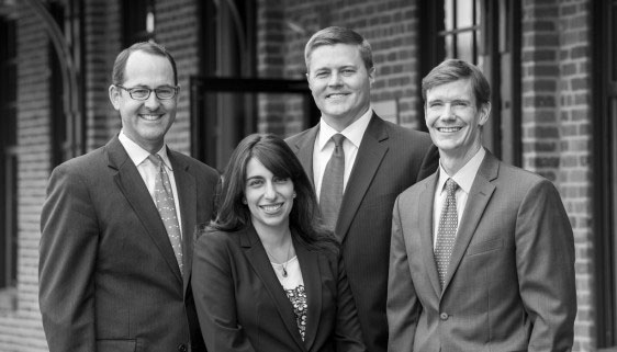 Four people in business attire stand together outdoors in front of a brick building, smiling at the camera. The photo is black and white.