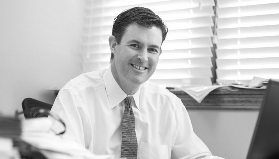 A man in a dress shirt and tie sits at a desk, smiling at the camera. There are papers and a laptop on the desk, and blinds cover the window behind him. The image is in black and white.